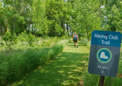 A green grassy trail winds through a lush, wooded area in Split Rock Creek State Park. A Hiking Club Trail sign marks the 2.6-mile path as two people with backpacks walk ahead along the scenic route.