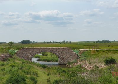 A stone arch bridge crosses a small creek amid grassy fields and rolling hills under a partly cloudy sky at Split Rock Creek State Park. Cyclists ride on a nearby path, while the scenic landscape stretches into the distance.
