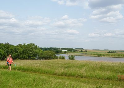 A person wearing an orange shirt and backpack walks along a grassy path near a river in Split Rock Creek State Park, with green trees, open fields, and a partly cloudy sky in the background.