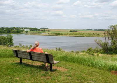 A person sits alone on a bench facing a calm lake at Split Rock Creek State Park, surrounded by green grass and trees, under a partly cloudy sky. The rural landscape features open fields stretching into the distance.