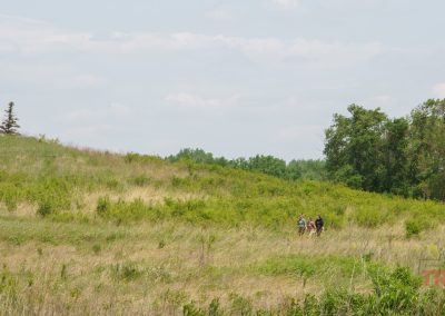 A group of three people walk along a grassy trail through the rolling green hills of Split Rock Creek State Park under a partly cloudy sky, with trees in the background.
