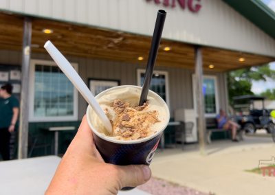 A hand holds up a cup of Pipestone Dari King Glacier ice cream topped with crushed candy and two straws in front of a small building with a porch. People and bicycles are visible in the background.