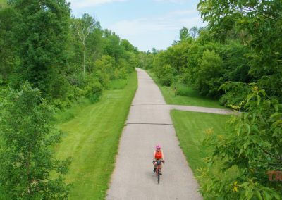 A person wearing a helmet and orange shirt rides a bicycle on a paved trail surrounded by lush green trees and grass under a blue sky.