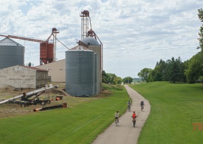 Four cyclists ride on a paved path past large grain silos and industrial buildings, with green grass and trees lining the path under a partly cloudy sky.