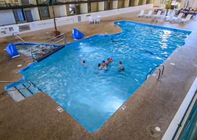 Indoor pool area with several people swimming. There is a small hot tub to the left, surrounded by chairs and tables. The pool is bordered by tan flooring, and the space is well-lit with seating nearby.
