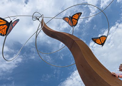 A woman looks up at a tall sculpture of monarch butterflies and swirling metal against a bright, cloudy sky. The butterflies appear to be flying above the curving, rust-colored structure.