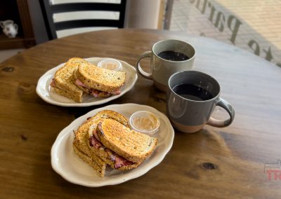 Two plates with stacked sandwiches and small containers of sauce, along with two mugs of black coffee, are set on a round wooden table by a window.