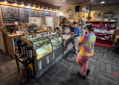 A woman in a tie-dye shirt waits at a coffee shop counter while a man orders. The shop has a chalkboard menu, a glass pastry display, a souvenir shelf, and people sitting at tables.