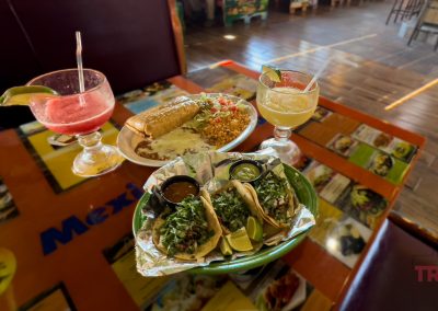 A table with three tacos topped with cilantro and onions, lime wedges, a plate with an enchilada, rice, and salad, and two colorful margarita drinks garnished with lime, inside a Mexican restaurant.