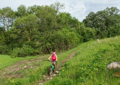 A person in a pink outfit and helmet rides a mountain bike on a narrow dirt trail through a green, grassy area with trees and shrubs under a partly cloudy sky.