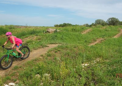 A person wearing a pink helmet and shirt rides a green mountain bike on a winding dirt trail through a grassy, green landscape under a partly cloudy sky.