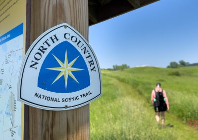 A sign for the North Country National Scenic Trail is mounted on a post next to a trail map. In the background, a person walks along a grassy path through open fields under a clear blue sky.