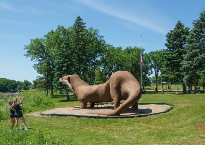 Two people take a selfie in front of a large, brown otter sculpture in a grassy park with trees and a small lake in the background on a sunny day.