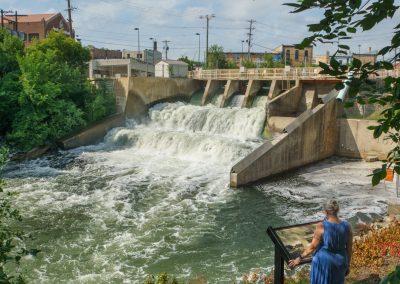 A woman in a blue dress stands on a platform, overlooking a concrete dam with rushing water and surrounding greenery, in an urban area with buildings and power lines in the background.