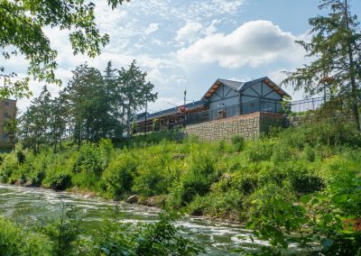 A modern building with a patio sits atop a stone wall, overlooking a lush green riverside with trees and bushes under a partly cloudy sky.