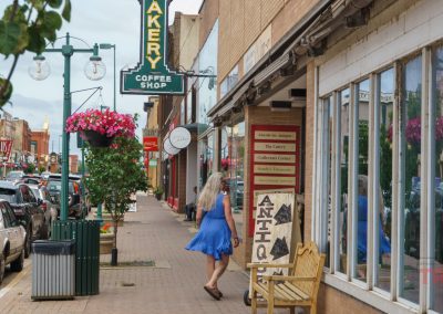 A woman in a blue dress walks down a small-town sidewalk lined with shops, including a bakery and antique store, on a cloudy day. Flowers hang from street lamps and parked cars line the street.