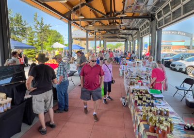 People shop at an outdoor market with tables displaying various goods like jars and bags. Some shoppers browse while others walk along the covered area. Cars and trees are visible in the background. A sign reads WATERFRONT PARK.