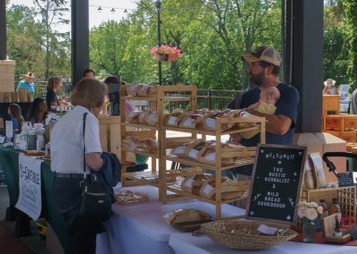 A man holding a baby stands behind a table of baked goods at an outdoor market, talking to a woman customer. Other vendors and shoppers are visible in the background under a shaded pavilion.