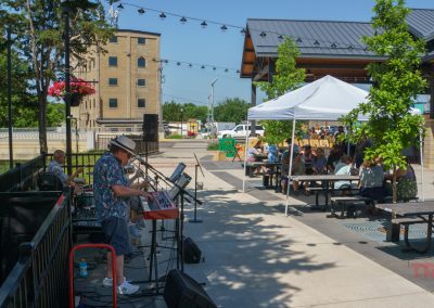 A small band performs outdoors under string lights beside a river while people sit at tables under a white canopy, enjoying the sunny day at a community event space.