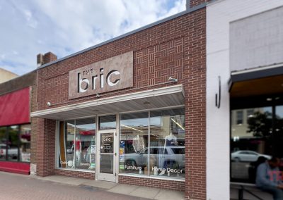 Street view of a store called The Bric with large front windows displaying various items. The sign mentions jewelry, furniture, and home decor. The building has a brown brick facade and is located between other storefronts.