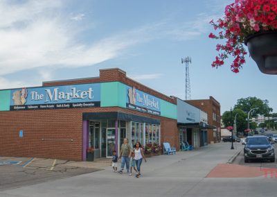 A brick storefront with a blue sign reading The Market sits on a small-town street. Three people walk on the sidewalk, flowers hang in a basket overhead, and parked cars line the street on a cloudy day.