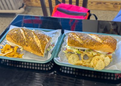 Two trays on a black table, each with a sub sandwich on a seeded roll, filled with lettuce, tomato, and other toppings, served with chips. A pink bag and a persons arm are in the background.