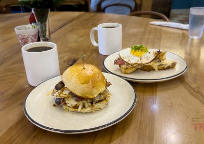 Two plates of breakfast food on a round wooden table: one with a sandwich of eggs, mushrooms, and cheese on a bun, the other with toast, ham, and a fried egg. Two white mugs of coffee and a glass of water are also on the table.