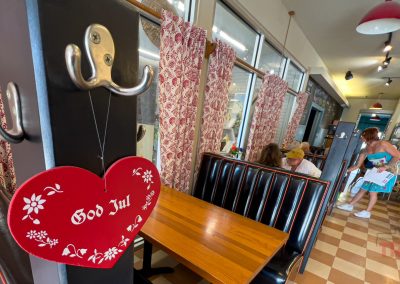 A red heart-shaped sign reading God Jul hangs on a coat rack in a cozy café with checkered floors, wooden tables, and people dining and chatting in the background.