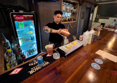 A bartender prepares a drink behind a wooden bar, with various bottles, a fridge, and mixers visible. The bar is neatly organized, and the atmosphere appears casual and inviting.