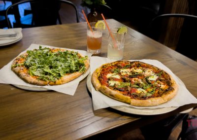 Two pizzas sit on a wooden table, one topped with fresh arugula and the other with various vegetables. Two cocktails with straws and garnishes are beside them. Plates, napkins, and a glass of water are in the background.