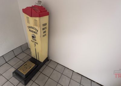 A vintage fortune-telling machine with a red and yellow top stands in a tiled corner, offering Character Readings and to tell Your "Wate" and Fate for 2 cents.