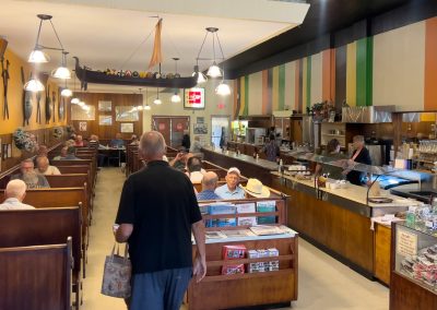 A retro-style diner with wooden booths, customers seated and eating, and a counter on the right where staff are serving food. Colorful stripes decorate the wall above the counter, and various decor items are displayed throughout.