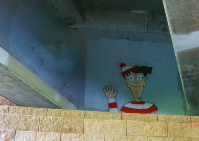 A mural of the character Waldo, wearing his red and white striped shirt, hat, and glasses, is painted on a concrete wall under a bridge. He is waving and partially obscured by a tan brick wall.
