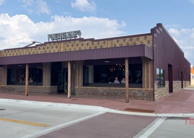 A brick and metal building with large windows and a sign on top reading Forbidden Barrel Brewing. The building sits on a clean, wide sidewalk under a partly cloudy sky.