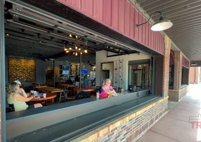 People sit at tables inside a restaurant near a large open window, with a few diners visible. The restaurant has exposed ceilings, warm lighting, and brick details.