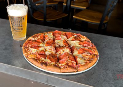 A glass of beer and a pepperoni pizza on a gray countertop in a restaurant setting, with wooden chairs in the background.