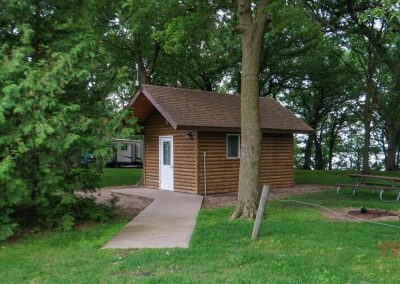 A small wooden cabin sits among tall trees in a grassy park, with a sidewalk leading to its door. A picnic table and fire pit are nearby, and a camper is visible in the background.