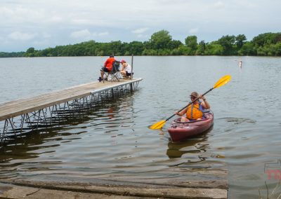 A person paddles a red kayak near a dock, where three people sit facing the lake. Trees line the far shore under a cloudy sky. The water is calm.