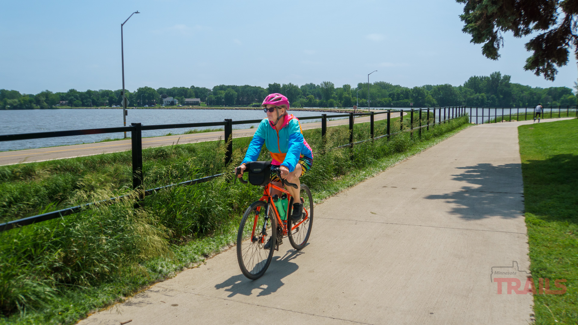 Okabena Lake Trail A person wearing a pink helmet and colorful jacket rides a bicycle on a paved trail beside a lake, bordered by grass and a black metal fence, under a clear sky.