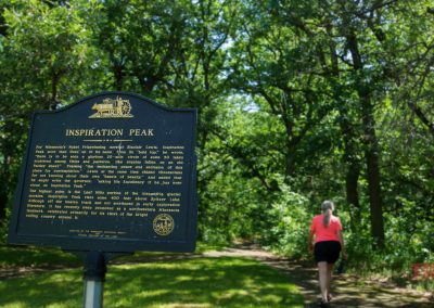 A black and gold historical marker for Inspiration Peak stands on a leafy trail, with a woman in a pink top and black shorts walking away on the sunlit wooded path.