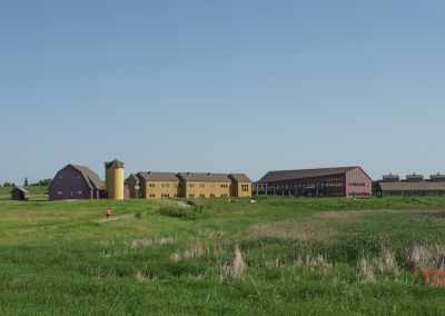 A rural landscape with green grass and a large, barn-like building complex in yellow, red, and purple. A small pond and a person in red walking are visible under a clear blue sky.