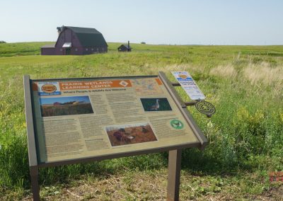 An informational sign for the Prairie Wetlands Learning Center stands in a grassy field, with a large barn-like building in the background under a clear sky.