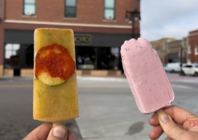 Two hands holding popsicles outdoors: one is yellow-green with a red chili circle, the other is pink with dark specks. The background shows brick buildings and a street scene.