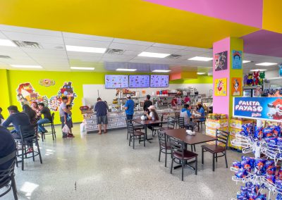 A colorful, lively ice cream shop with bright yellow and pink walls, customers sitting at tables and the counter, and various candy displays and cartoon-themed wall decorations.