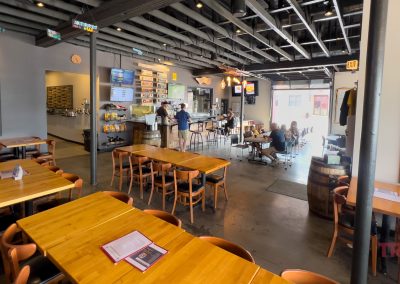 A spacious, modern restaurant interior with wooden tables and chairs, a central bar area, people dining, wall-mounted TVs, and exposed ceiling beams, creating a relaxed and welcoming atmosphere.
