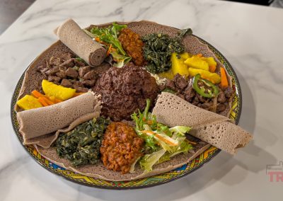 A colorful platter of Ethiopian food featuring injera rolls, various stewed vegetables, lentils, greens, salad, and a spicy meat stew arranged on a decorative plate atop a marble surface.