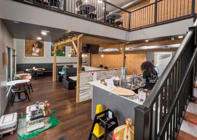 A cozy, modern café with wood flooring, a mezzanine seating area, black booths, and a counter where a barista prepares drinks. Decorative items and art add warmth to the inviting atmosphere.