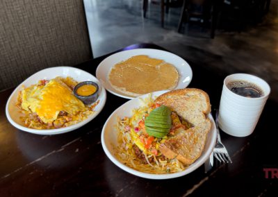 A table with three breakfast dishes: a cheesy hash brown bowl with sauce, pancakes, and a veggie-topped egg bowl with toast and avocado. A cup of black coffee sits nearby.