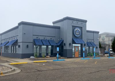 A gray and blue restaurant building with blue awnings stands in an empty parking lot. Several handicap parking spaces are near the entrance. The sign above the door reads THE MAV GRILL. The sky is overcast.