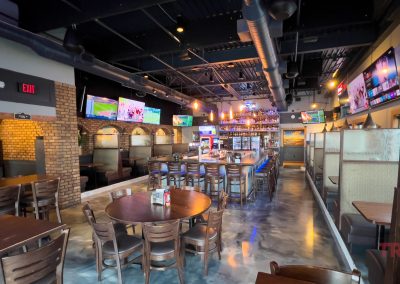 A modern sports bar with wooden tables, a central bar area, multiple TV screens displaying sports, and booth seating along the walls. The atmosphere is vibrant and welcoming.